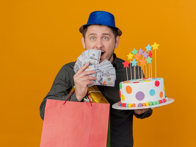 Surprised young party guy wearing blue hat holding gifts with cake and covered face with cash isolated on orange