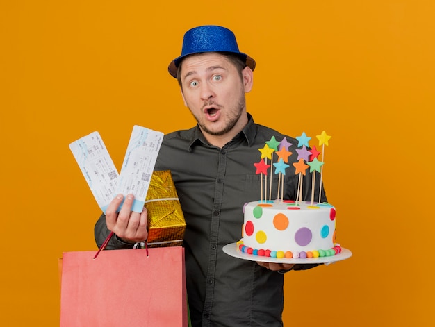 Surprised young party guy wearing blue hat holding gifts with cake and tickets isolated on orange