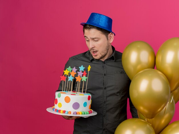 Удивленный молодой тусовщик в синей шляпе, стоящий рядом с воздушными шарами, держащий и смотрящий на торт, изолированный на розовом