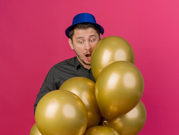 Surprised young party guy wearing blue hat standing behind balloons isolated on pink