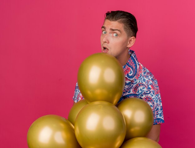 Surprised young party guy wearing colorful shirt standing behind balloons isolated on pink
