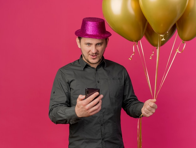 Surprised young party guy wearing pink hat holding balloons and holding and looking at phone isolated on pink