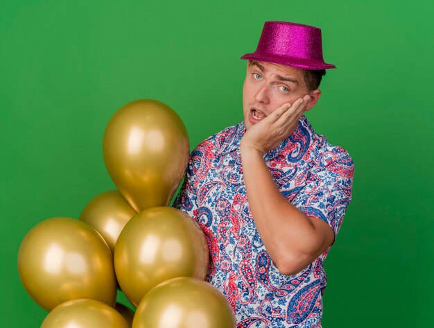 Surprised young party guy wearing pink hat holding balloons putting hand on cheek isolated on green