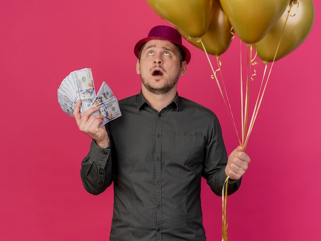 Free photo surprised young party guy  wearing pink hat holding balloons with cash isolated on pink