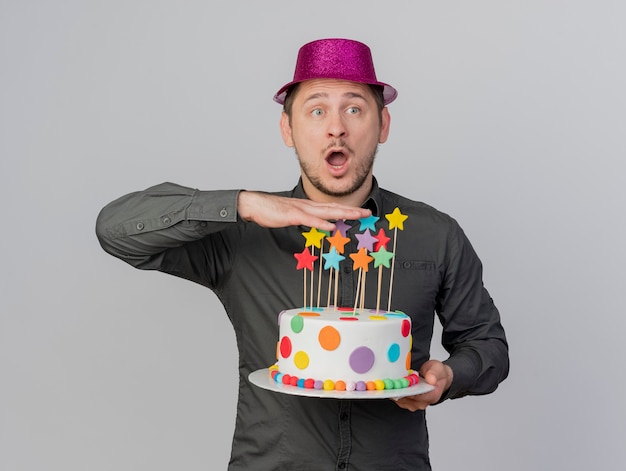 Surprised young party guy wearing pink hat holding cake isolated on white background