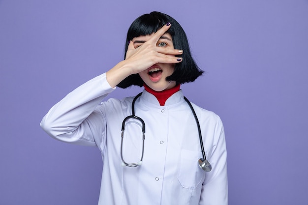 Surprised young pretty caucasian girl in doctor uniform with stethoscope covering her face with hand and looking at front through fingers