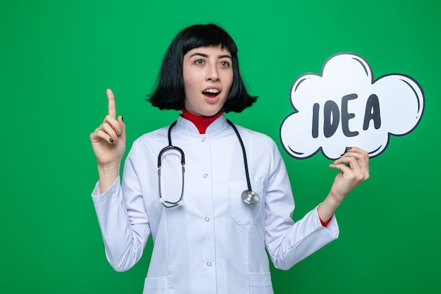 Surprised young pretty caucasian girl in doctor uniform with stethoscope looking at side pointing up and holding idea bubble 
