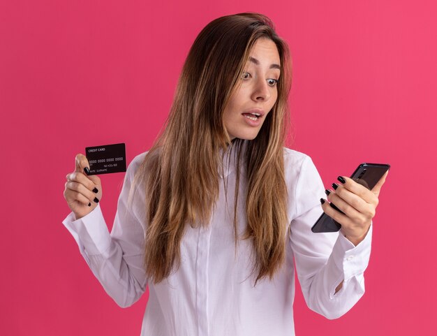 놀란 된 젊은 예쁜 백인 여자 신용 카드를 보유하고 복사 공간이 분홍색 벽에 고립 된 전화에서 보이는
