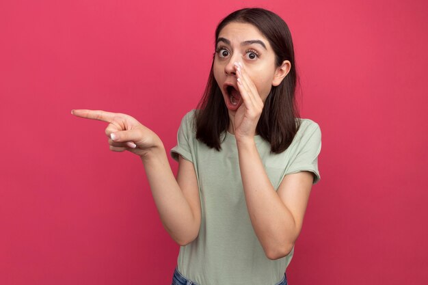 Surprised young pretty caucasian girl  pointing at side keeping hand near mouth whispering isolated on pink wall with copy space