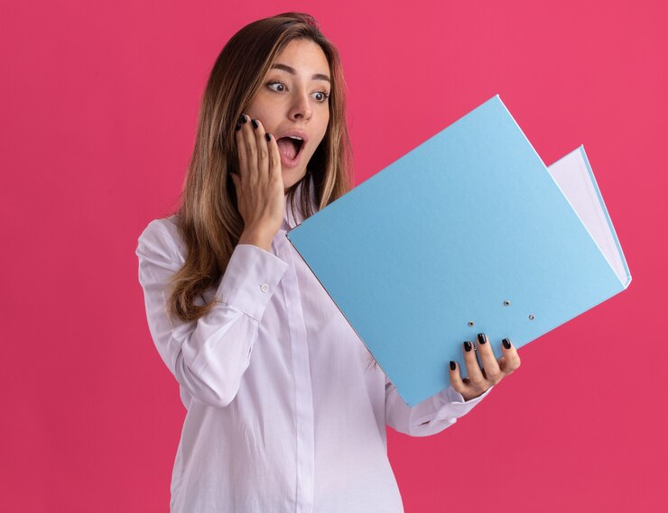 मुफ़्त फोटो surprised young pretty caucasian girl puts hand on face holding and looking at file folder on pink 