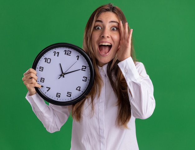 Free photo surprised young pretty caucasian girl puts hand on face and holds clock on green