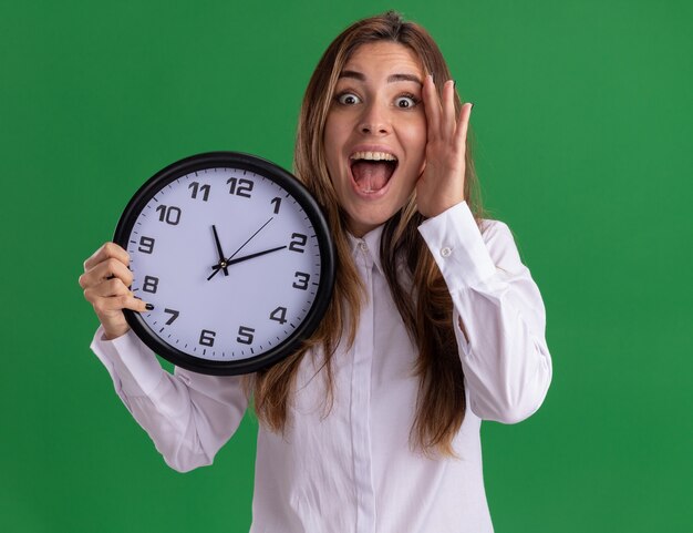 Surprised young pretty caucasian girl puts hand on face and holds clock on green 