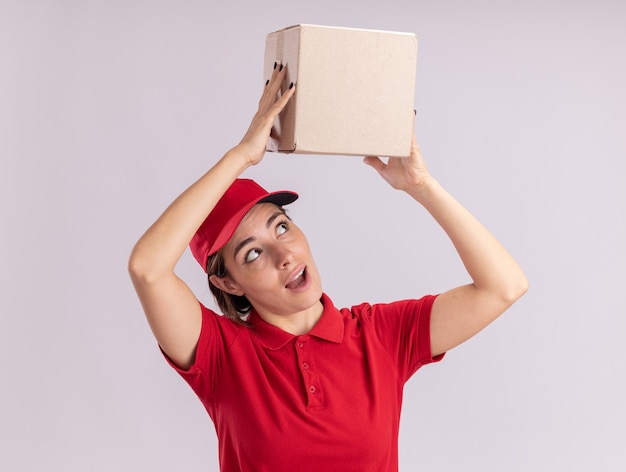 Surprised young pretty delivery girl in uniform holds cardbox over head on white