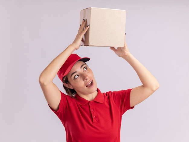 Удивленная молодая симпатичная доставщица в униформе держит картонную коробку над головой на белом