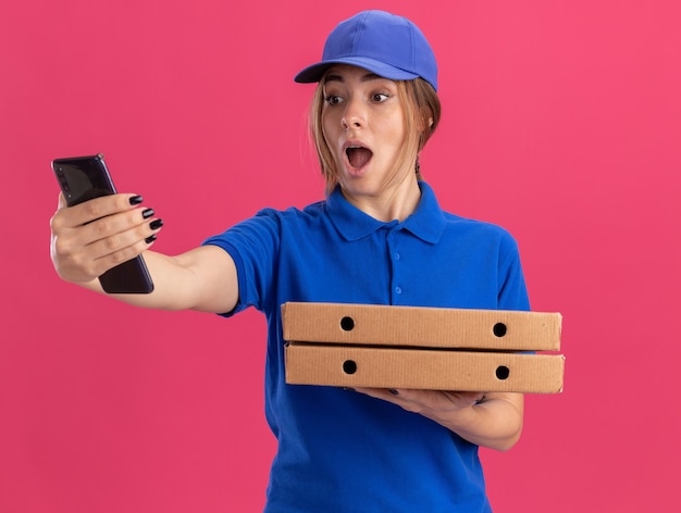 La giovane ragazza graziosa di consegna sorpresa in uniforme tiene le scatole della pizza e guarda il telefono sul rosa