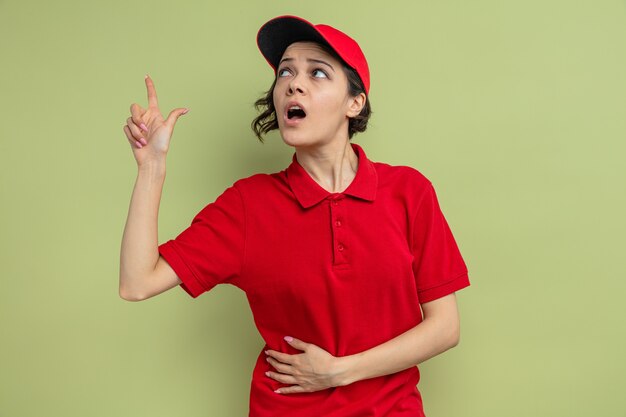 Surprised young pretty delivery woman looking and pointing up 