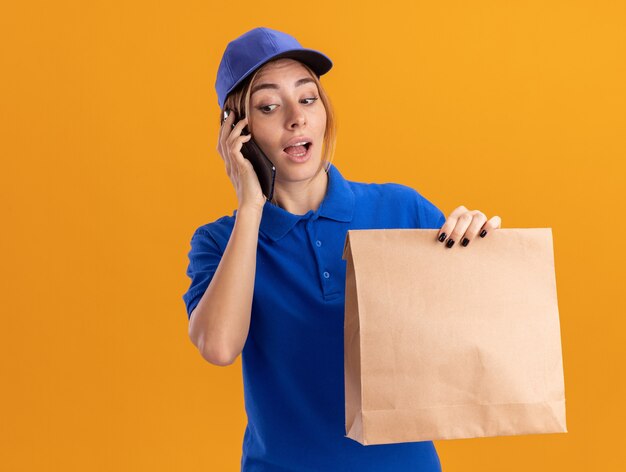 Surprised young pretty delivery woman in uniform holds paper package and talks on phone isolated on orange wall