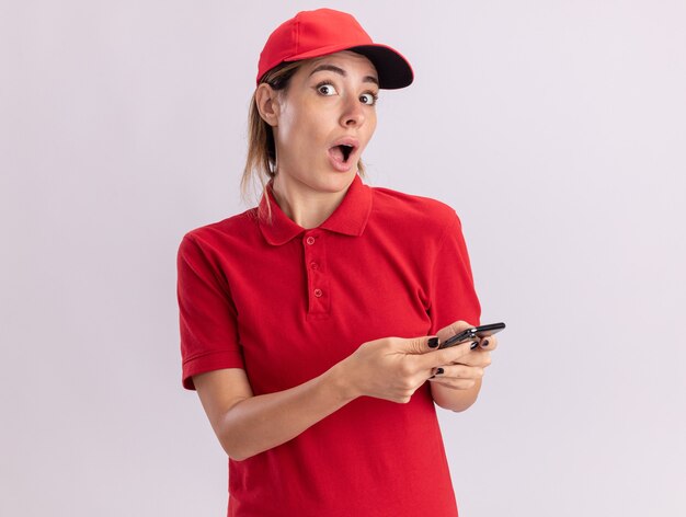 Surprised young pretty delivery woman in uniform holds phone isolated on white wall