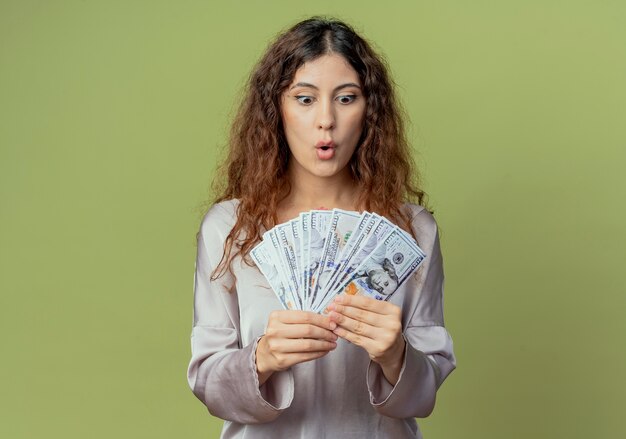 Surprised young pretty female office worker holding and looking at cash isolated on olive green