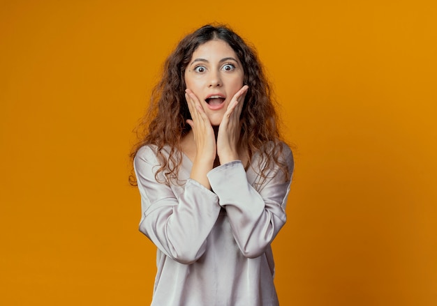 Surprised young pretty girl putting hands on cheeks isolated on yellow wall