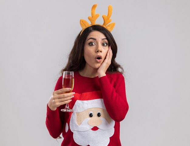 Surprised young pretty girl wearing reindeer antlers headband and santa claus sweater holding glass of champagne looking keeping hand on face 