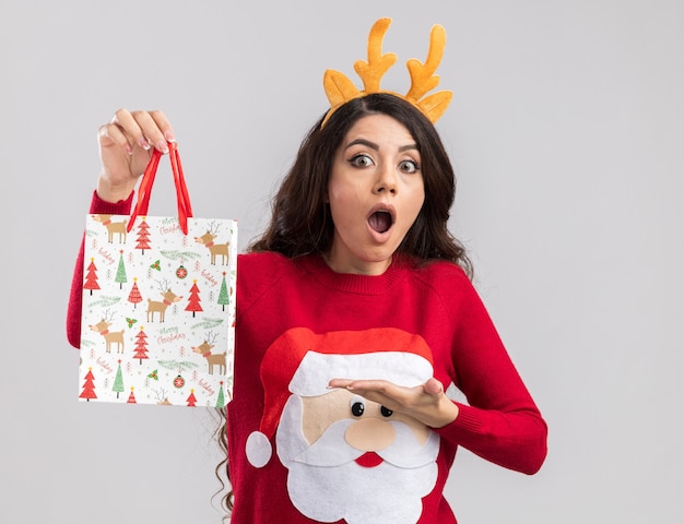 Surprised young pretty girl wearing reindeer antlers headband and santa claus sweater holding and pointing at christmas gift bag looking 