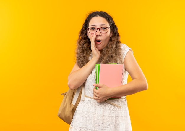 Surprised young pretty schoolgirl wearing glasses and back bag holding book and note pad putting hand near mouth isolated on yellow wall