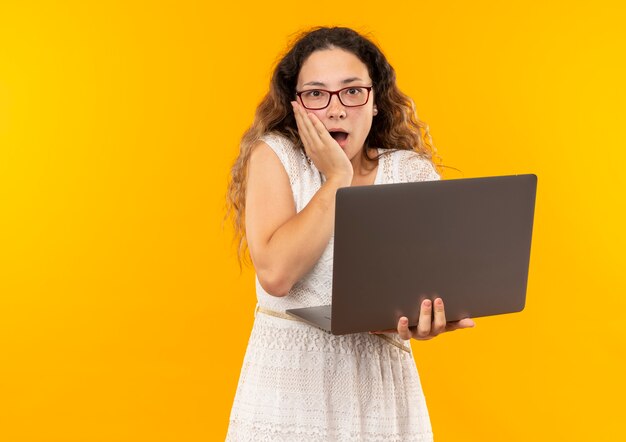 Surprised young pretty schoolgirl wearing glasses and back bag holding laptop with hand on face isolated on yellow wall