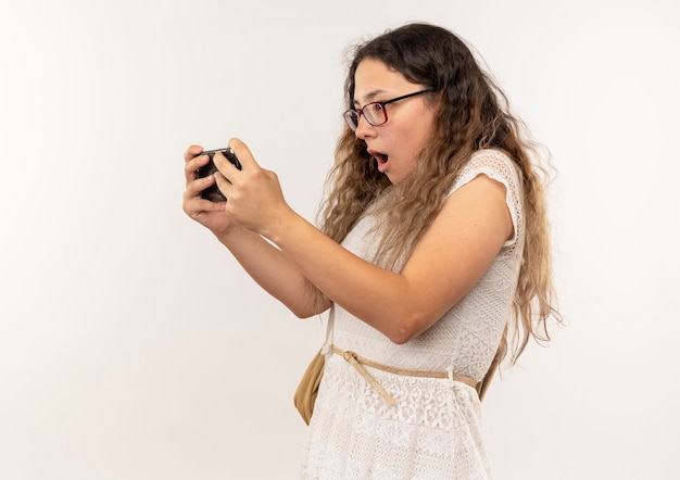 Sorpreso giovane studentessa graziosa con gli occhiali e borsa posteriore utilizzando il suo telefono isolato sulla parete