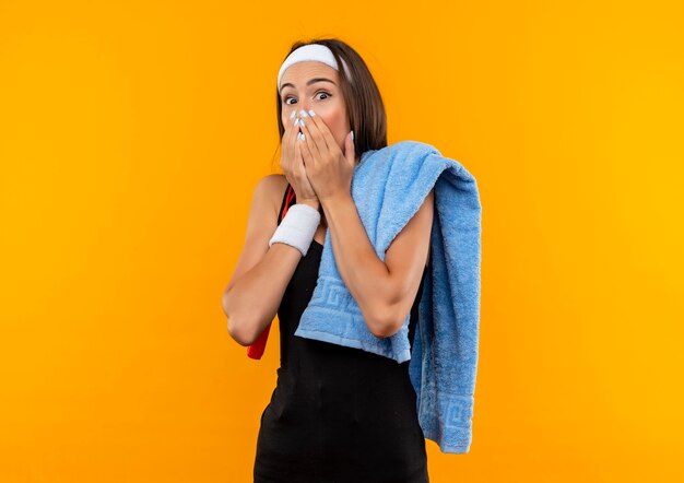 Surprised young pretty sporty girl wearing headband and wristband with towel and jumping rope on shoulders putting hands on mouth isolated on orange space 