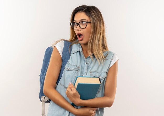 眼鏡とバックバッグを身に着けて本を保持し、白い壁に隔離された側を見て驚いた若いかわいい学生の女の子
