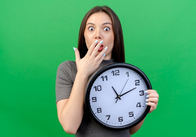 Surprised young pretty woman holding clock putting hand on mouth isolated on green background with copy space