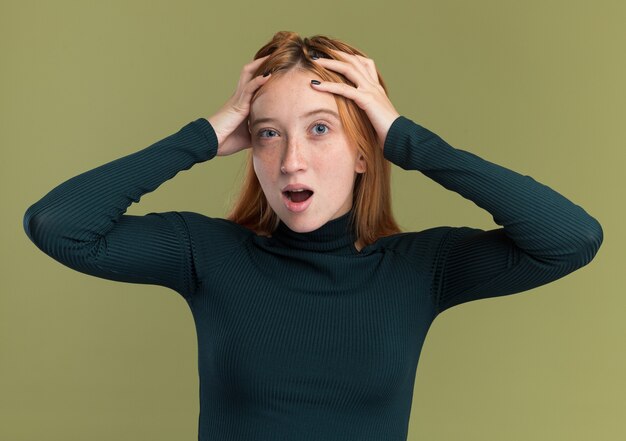 surprised young redhead ginger girl with freckles puts hands on head on olive green