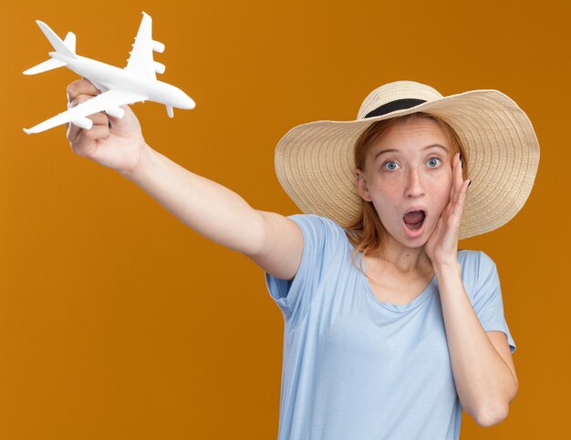 Surprised young redhead ginger girl with freckles wearing beach hat holds model plane isolated on orange wall with copy space