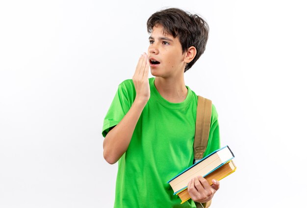 Surprised young school boy wearing backpack holding books covered mouth with hand isolated on white wall