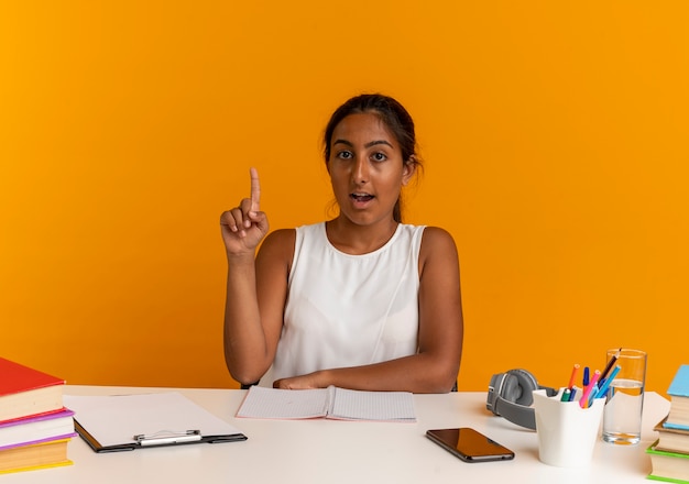 Surprised young schoolgirl sitting at desk with school tools points at up on orange