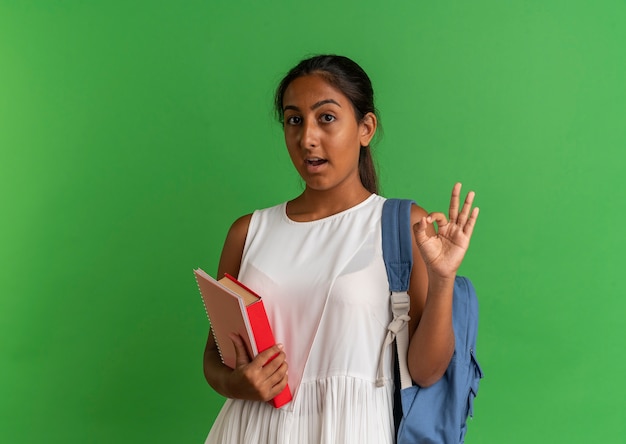 Surprised young schoolgirl wearing back bag holding book with notebook and showing okey gesture on green