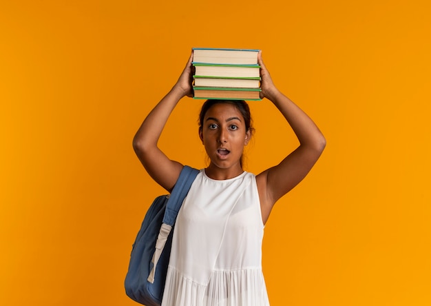 Surprised young schoolgirl wearing back bag holding books on head on orange