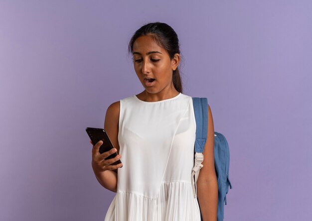 Surprised young schoolgirl wearing back bag holding and looking at phone on purple