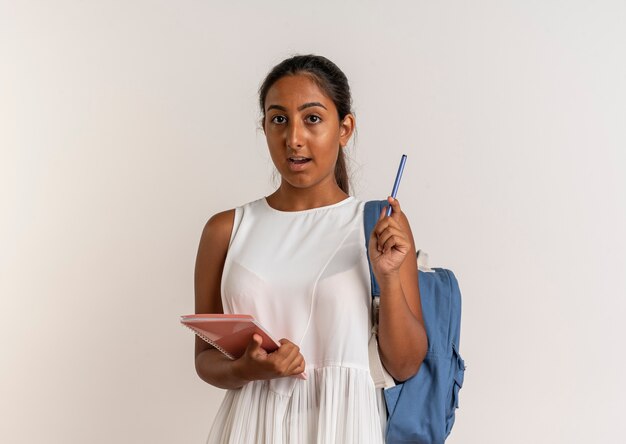 Surprised young schoolgirl wearing back bag holding notebook and pen on white