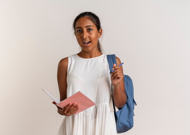 Удивленная молодая школьница в задней сумке, держащая блокнот и ручку на белом
