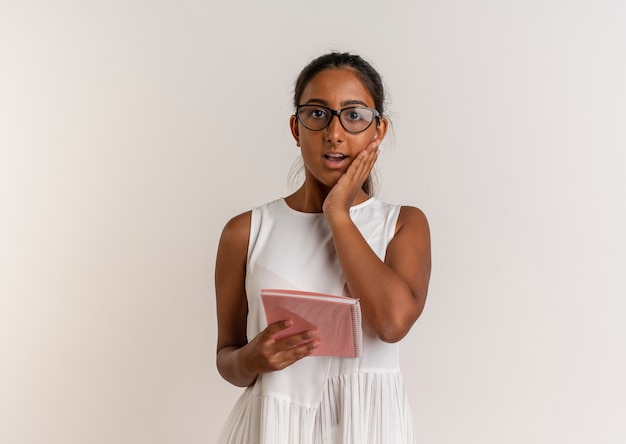Surprised young schoolgirl wearing glasses putting hand on cheek isolated on white wall