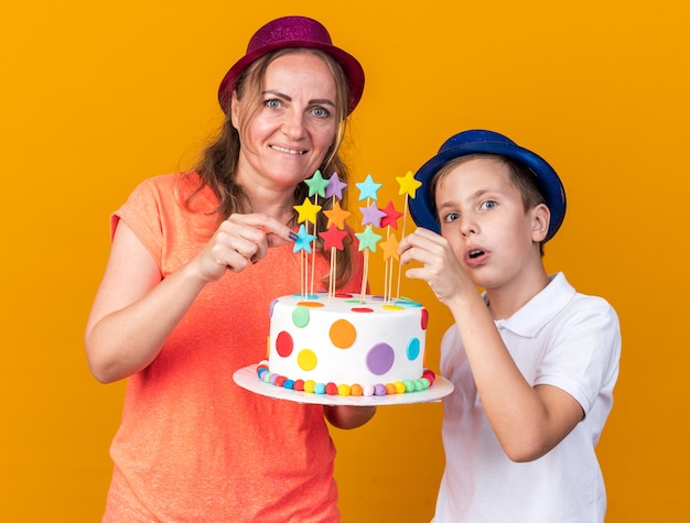 Sorpreso giovane ragazzo slavo con cappello da festa blu che tiene la torta di compleanno con sua madre che indossa un cappello da festa viola isolato sulla parete arancione con spazio per la copia