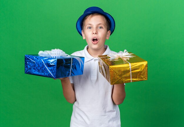 удивленный молодой славянский мальчик в синей шляпе, держащий подарочную коробку в каждой руке, изолированной на зеленой стене с копией пространства