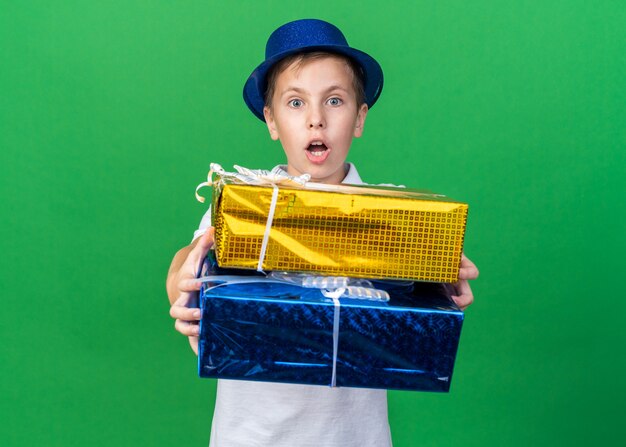 コピースペースで緑の壁に分離されたギフトボックスを保持している青いパーティーハットで驚いた若いスラブ少年