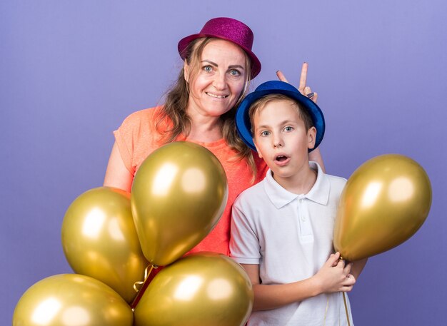 surprised young slavic boy with blue party hat holding helium balloons with his mother wearing violet party hat isolated on purple wall with copy space