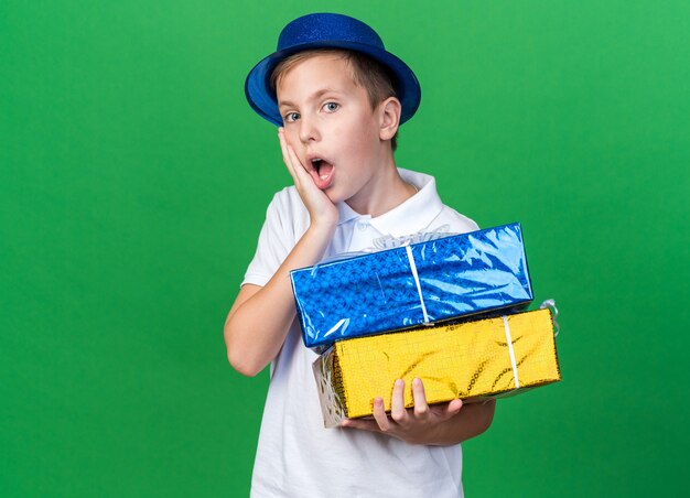 surprised young slavic boy with blue party hat putting hand on face and holding gift boxes isolated on green wall with copy space