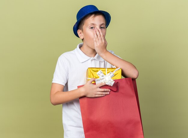 surprised young slavic boy with blue party hat putting hand on mouth and holding gift box in shopping bag isolated on olive green wall with copy space