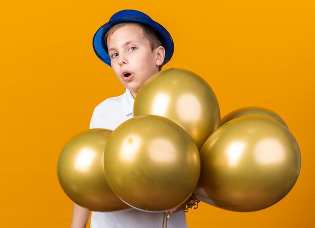 コピースペースとオレンジ色の壁に分離されたヘリウム気球と立っている青いパーティーハットで驚いた若いスラブ少年