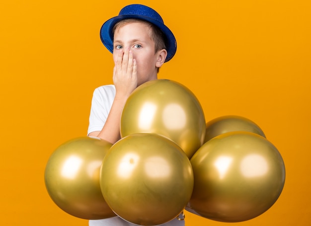 удивленный молодой славянский мальчик в синей партийной шляпе, стоящий с гелиевыми шарами, положив руку на рот, изолированный на оранжевой стене с копией пространства